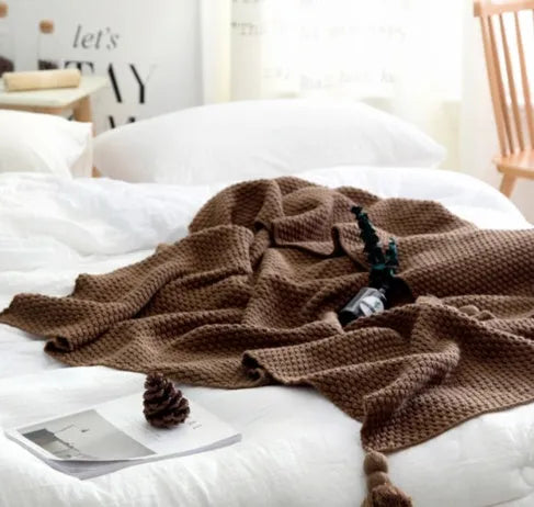 Nordic style fringed knit throw blanket in a neutral colour, shown draped over a sofa.