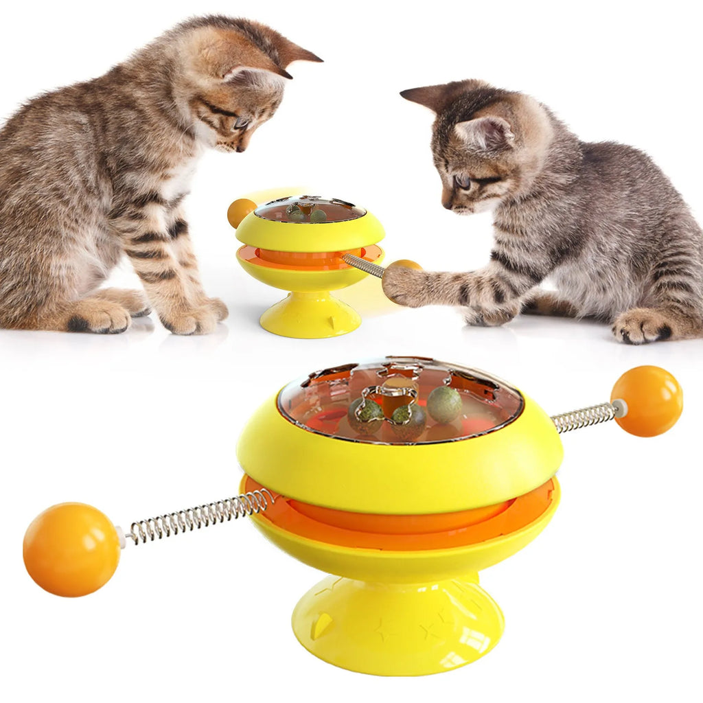 Rotating turntable cat toy with colourful base and transparent top containing three catnip balls, on a wooden floor.