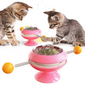 Rotating turntable cat toy with colourful base and transparent top containing three catnip balls, on a wooden floor.