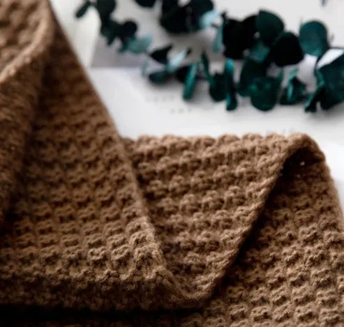 Nordic style fringed knit throw blanket in a neutral colour, shown draped over a sofa.
