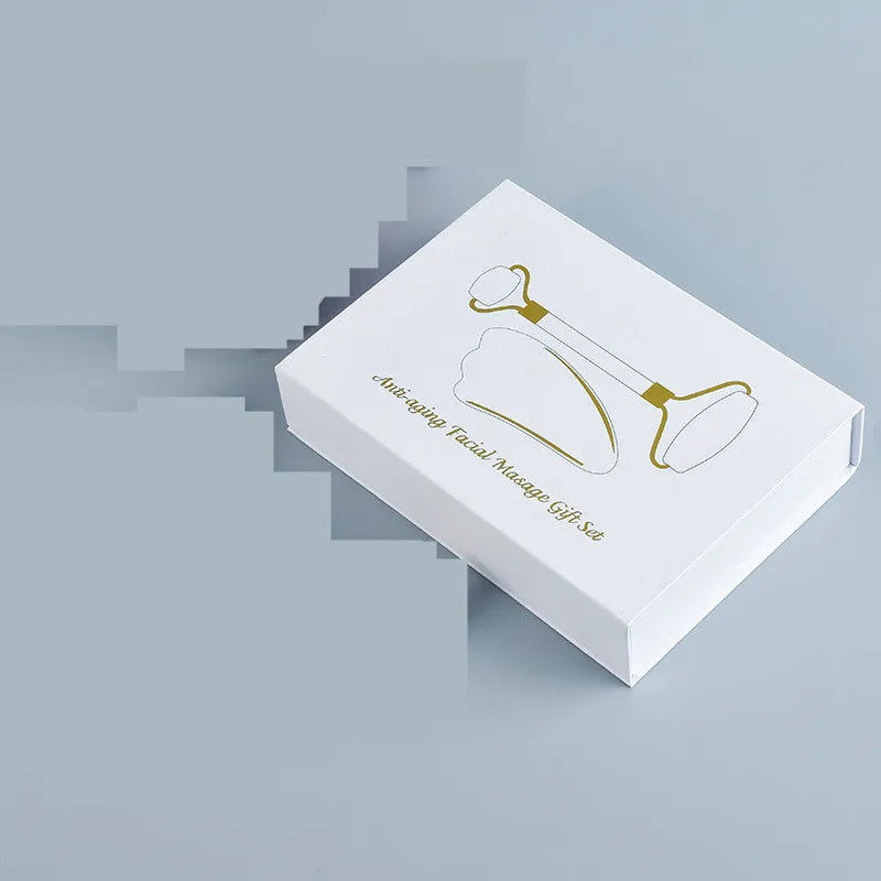 White jade facial roller and gua sha board set on a plain surface.
