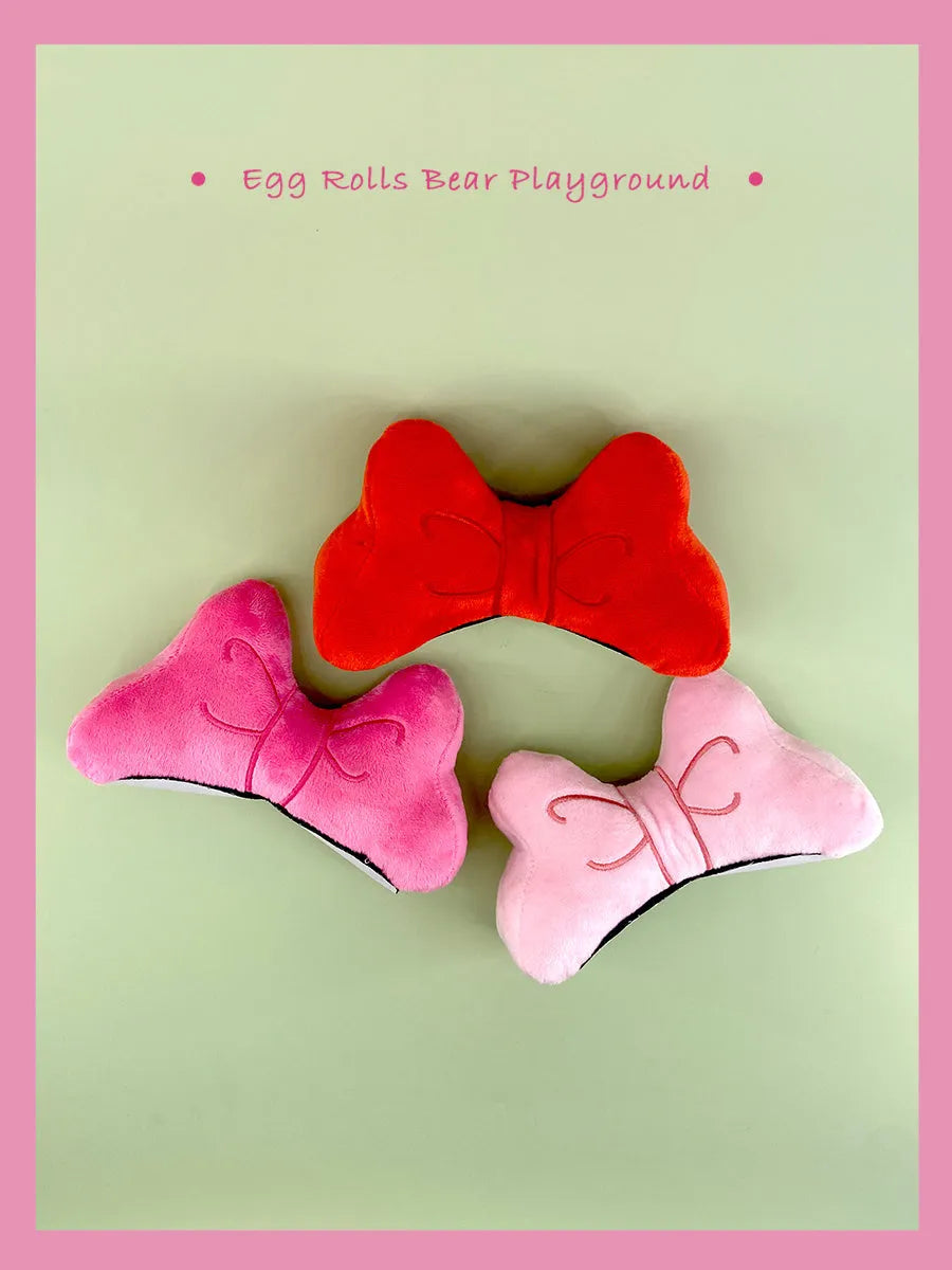 A soft plush bow tie helmet decoration shown in red, peach pink, and rose pink colours.