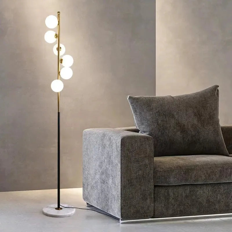 A tall, modern minimalist floor lamp with a wrought iron and glass body standing on a circular marble base.