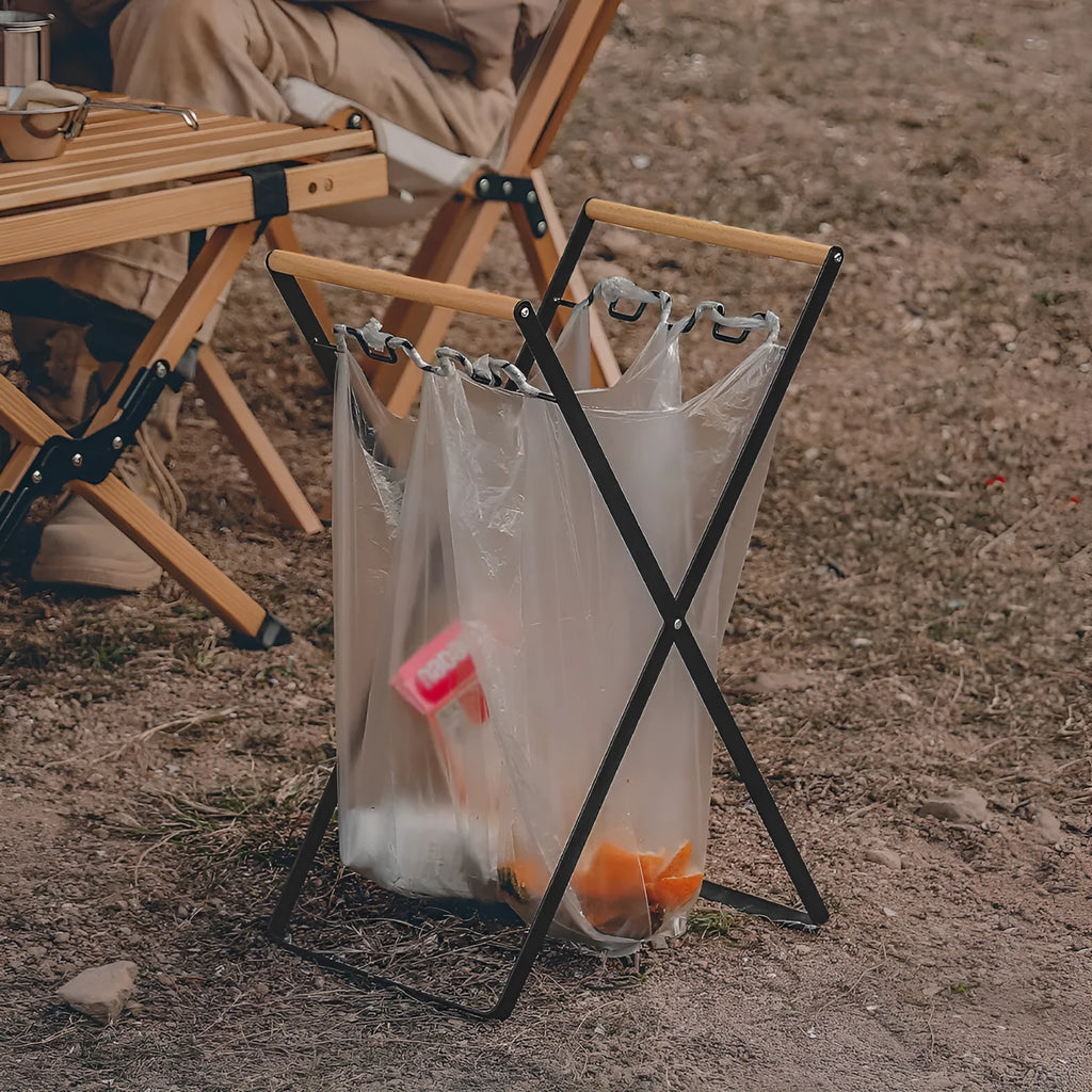Black foldable iron trash storage hanger with wooden handle holding a bin bag outdoors.